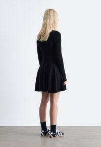 Schwarzes, geripptes Strickkleid mit langen Ärmeln, tailliertem Oberteil und schwingendem Rock. Getragen mit schwarz-weißen Sportschuhen und dunkelblauen Socken.