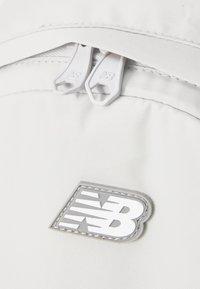 Hvit stoff med en glatt tekstur, med to hvite glidelåsdragere. Har en sølv- og grå New Balance-logo på en brodert lapp.