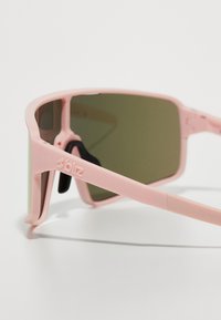 Ochelari de soare cu un cadru din plastic roz deschis, lentile verzi și accente negre. Logo vizibil pe lateral și un design unghiular distinct.