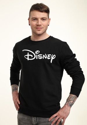 Junger Mann mit kurzen braunen Haaren, der einen schwarzen Disney-Sweatshirt trägt, steht mit den Händen in der Hüfte vor einem weißen Hintergrund.