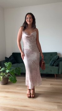 Mladá žena v ľahkých šatách so zvieracím vzorom a hnedými platformovými lodičkami stojí vo vnútri a drží hnedú kabelku pri zelenej pohovke a rastline.