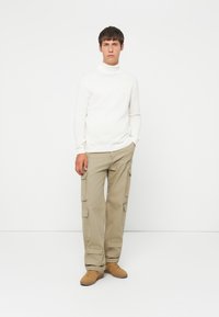 Jersey de cuello alto blanco, pantalones cargo beige claro con bolsillos laterales y zapatillas color tostado, sobre un fondo neutral.