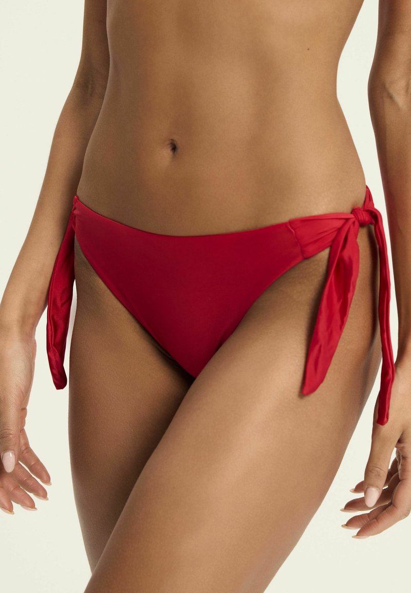 Czerwone majtki bikini wykonane z gładkiego materiału, z bocznymi trokami i niskim stanem. Tekstura wydaje się miękka i elastyczna.