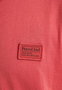 Rode stof met een gladde textuur heeft een rechthoekig zwart label erop gestikt, met de tekst "Petrol Ind" en aanvullende merkdetails.