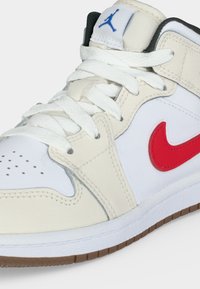 Υψηλό λευκό και μπεζ αθλητικό παπούτσι με κόκκινο λογότυπο Nike, λευκά κορδόνια, διάτρητη μύτη και μπλε λογότυπο Jumpman στη γλώσσα.