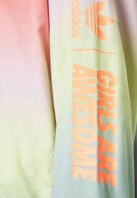 Adidasjacka med en gradientdesign som går från rosa till grön, med orange text som säger "GIRLS ARE AWESOME" och logotyp på ärmen.