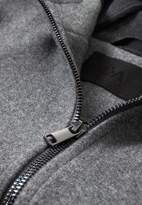 Gros plan sur une veste en polaire grise avec une fermeture éclair en métal argenté partiellement ouverte et une étiquette en tissu noir à l'intérieur du col.