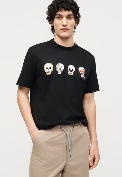 Νέος άνδρας που φοράει μαύρο μπλουζάκι με τέσσερα πολύχρωμα σχέδια κρανίων στο στήθος και μπεζ παντελόνι με κορδόνι.