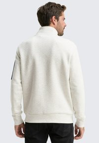 Witte gewatteerde sweatshirt met een hoge kraag, textuurstof en ribgebreide manchetten. Zwarte accent op de linkermouw en donkere jeans worden gedragen.