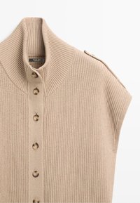 Beige knitted sleeveless cardigan with high ribbed collar and front buttons, featuring a shoulder strap detail on one side.