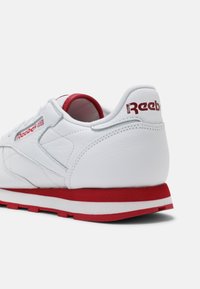 Baskets en cuir blanc avec un onglet de talon rouge et une semelle rouge. Logo "Reebok" imprimé en rouge à l'arrière et sur le côté. Texture lisse, design sportif.
