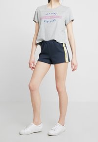 Graues Baumwoll-T-Shirt mit pinkem und blauem "Abercrombie New York"-Schriftzug, kombiniert mit Marineblauen Shorts mit gelben Seitenstreifen und weißen Sneakers.