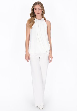 Młoda kobieta z długimi falującymi włosami, ubrana w białą, bezrękawową bluzkę na szyję i szerokie białe spodnie, stojąca na tle białego tła.