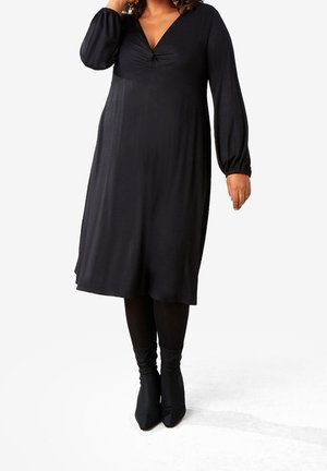 Vrouw draagt een zwarte midi-jurk met lange mouwen en een gerimpelde V-hals, gecombineerd met zwarte panty's en zwarte enkellaarsjes.