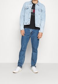 Světle modrá džínová bunda, černé tričko s grafikou a červenými a bílými pruhy, modré džíny a bílé tenisky; neformální vrstvený outfit.