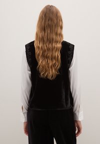 Gilet di velluto nero con ricami intricati sulle spalle, indossato sopra una camicetta bianca con polsini a bottoni; lunghi capelli ondulati e sciolti visibili.