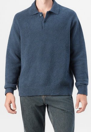 Mann trägt einen blauen, langärmeligen, strukturierten Polopullover und dunkelgraue Jeans vor einem einfarbigen Hintergrund.