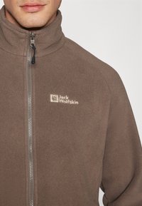 Smeđi flisani jakna s visokim ovratnikom, punim zatvaračem i izvezenim logom na grudima. Mekana tekstura i jednostavan dizajn.