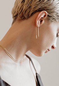 Des boucles d'oreilles dorées au design minimaliste pendent verticalement. Un délicat collier en or est porté, complétant un haut sans manches de couleur foncée.
