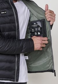 Crna puffer jakna sa srebrnim zatvaračem, koja se ističe glatkom vanjštinom, kviltanim uzorkom i zelenim unutrašnjim dijelom na kojem su prikazane oznake za njegu i ikone.