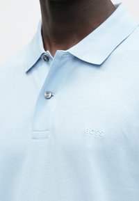 Tmavá osoba so svetlomodrou polokošeľou so zapnutými dvoma gombíkmi a vyšitým nápisom „BOSS“ na hrudi.