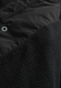 Sort jakke med en kombination af glat stof og tekstureret fleece. Bemærkelsesværdige detaljer inkluderer en trykknap og en kontrastfarvet stofsektion.