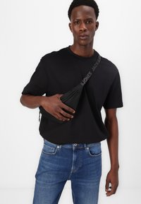 Black crew-neck t-shirt paired with blue denim jeans. The model holds a black sling bag with a printed strap. Simple and casual design.