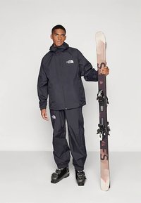 Mladý muž v černém lyžařském oblečení North Face držící svisle jednu lyži, má na sobě černé lyžařské boty, stojící na jednobarevném pozadí.