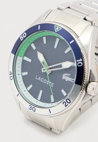 Sudraba nerūsējošā tērauda pulkstenis ar zilu un zaļu ierobežojumu, teksturēta pelēka ciparnīca, gaismu atstarotāji un Lacoste logs. Iekļauj modernu rokassprādzi.