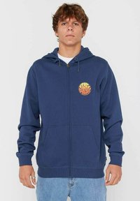 Jongeman in een marineblauwe zip-up hoodie met een rond oranje logo op de linkerborst en lichtblauwe spijkerbroek.