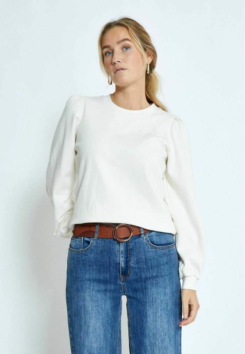 Felpa bianca a maniche lunghe con spalle a sbuffo, scollatura rotonda e tessuto testurizzato; abbinata a jeans blu e una cintura marrone.