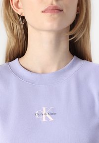 Svetlý levandulový rebrovaný sveter s okrúhlym výstrihom, ktorý na hrudi nesie biele logo Calvin Klein. Hladká textúra s priliehavým strihom.