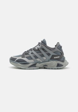 Zapatilla deportiva gris y negra con paneles de malla, superposiciones sintéticas en capas, cierre de cordones y suela acolchada con la marca "GUESS" en el talón.