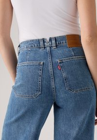 Achteraanzicht van een persoon in hoge taille blauwe spijkerbroeken met twee vierkante zakken en een bruine Levi's patch, gecombineerd met een witte mouwloze top.