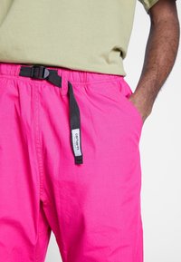 Klart rosa shorts i mjukt tyg, med ett svart justerbart bälte och sidofickor, tillsammans med en ljusgrön skjorta.