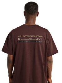 Homme portant un t-shirt marron avec un texte jaune "HIGH ALTITUDE LOW OPENING" et des lignes graphiques dans le haut du dos.