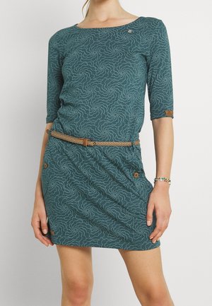 Mujer con un vestido color verde azulado con un patrón de puntos en espiral, mangas tres cuartos, un cinturón trenzado color beige y una pulsera de cuentas en una muñeca.