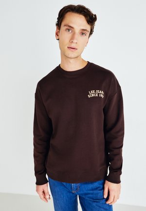 Brązowa bluza z okrągłym dekoltem z ribbingu, z złotą haftowaną naszkicowaną logo "LEE JEANS SINCE 1889" po lewej stronie klatki piersiowej. W połączeniu z niebieskimi dżinsami.