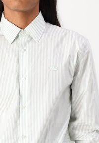 Šviesiai žalia ir balta dryžuota marškinėliai su mygtukais apykakle, turintys mažą Lacoste logotipą ant krūtinės ir lygią audinio tekstūrą.