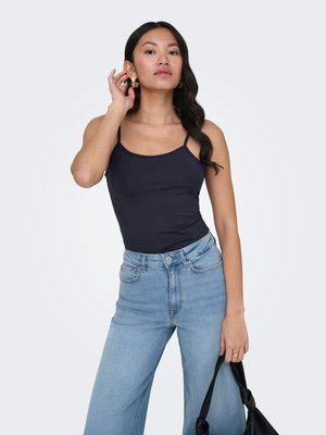 Woman wearing black tank top and light blue high-waist jeans, holding black handbag, adjusting gold hoop earring against plain background.