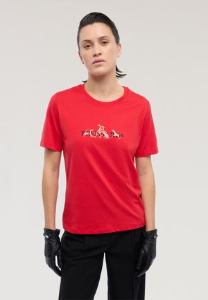 Vrouw met achterovergekamd haar draagt een rood grafisch T-shirt met de tekst "TRUST" en puppies, zwarte broek en zwarte leren handschoenen tegen een effen achtergrond.