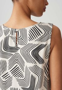Sleeveless blouse in cream with black wavy line pattern. Features a small button closure at the back neck and a smooth texture.