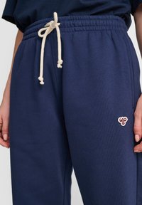 Pantaloni della tuta blu navy con vita elastica e cordino crema. Presentano un piccolo design ricamato sulla coscia sinistra. Tessuto morbido e testurizzato.