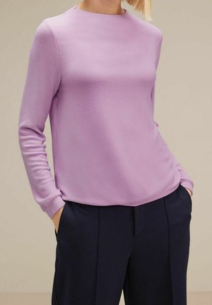 Persoon met een lichtpaarse top met lange mouwen, lichtjes ingestopt in donkerblauwe broek, met handen in de zakken tegen een effen achtergrond.