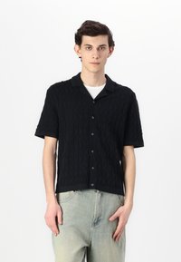 Chemise en tricot noire à manches courtes avec un motif de vagues texturé, un col avec boutons à l'avant, portée avec un pantalon de couleur claire.