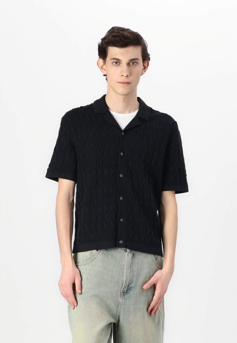 Schwarzes Kurzarm-Strickshirt mit strukturiertem Wellenmuster, einem Kragen und Frontknöpfen, kombiniert mit hellen Hose.