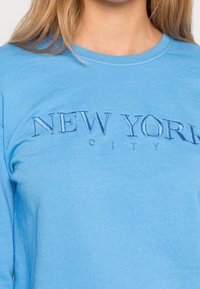 Svetle modré tričko s dlouhým rukávem, s tmavě modrým vyšitým nápisem "NEW YORK CITY" na hrudi, vyrobené z měkké bavlny.