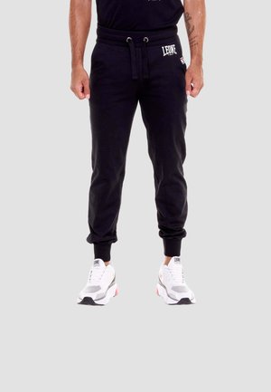 Uomo che indossa pantaloni da jogging neri con il logo "LEONE" sulla coscia e sneakers bianche, in piedi contro uno sfondo neutro.