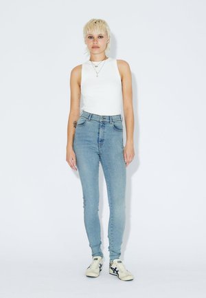 Person mit kurzen blonden Haaren, die ein weißes ärmelloses Oberteil, eine hellblaue hochgeschnittene Skinny-Jeans und weiße Turnschuhe trägt, steht vor einem einfarbigen Hintergrund.