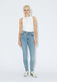 Persona con pelo corto rubio, vistiendo una blusa sin mangas blanca, jeans ajustados de cintura alta azul claro y zapatillas blancas, de pie contra un fondo liso.